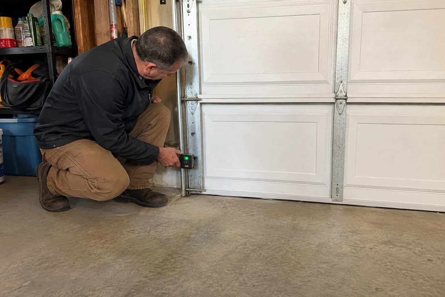 Technician kneeling beside a white paneled garage door, holding a small black box with a green light against the vertical track. — Photo-Eye Sensor Alignment or Replacement, Toronto